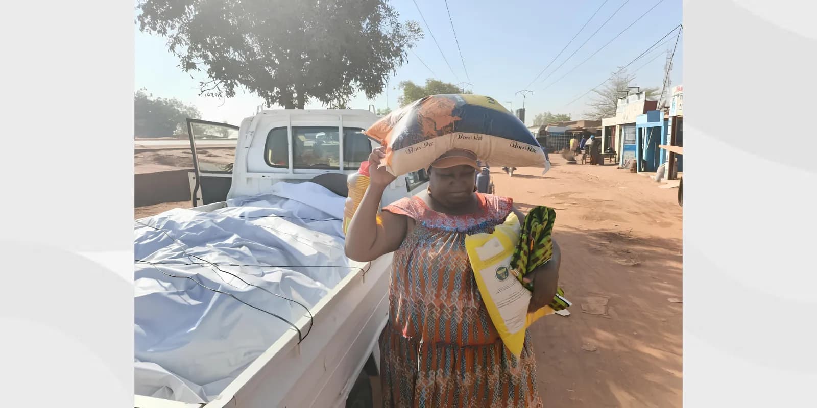 Une femme porte un sac de riz sur la tête devant un pick-up chargé de sacs, dans une rue